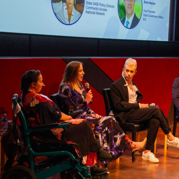 Panelists on stage at a rare disease policy discussion, one speaking into a microphone and one seated in a wheelchair.