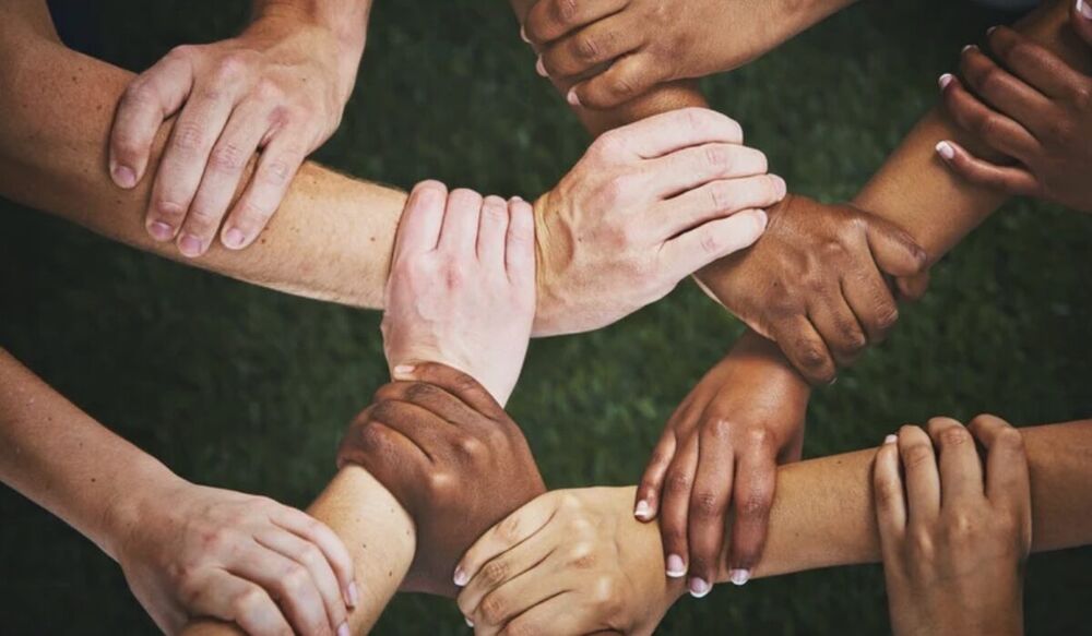 Hands and forearms of eight people clasped together in a ring over grass