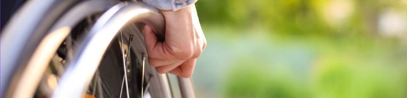 A close view of a person's hand resting on the push rim of their wheelchair, with greenery behind
